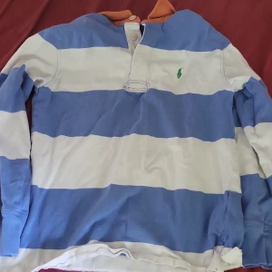 Randig pikétröja från Polo Ralph Lauren - Säljer en klassisk pikétröja från Polo Ralph Lauren med breda blå och vita ränder, lång ärm och orange krage. Tröjan har knappar vid halsen och den ikoniska gröna loggan broderad på bröstet. Perfekt för dig som gillar preppy stil. Pris kan diskuteras 