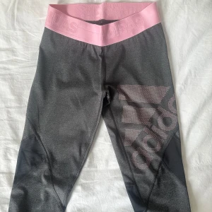 Grå och rosa Adidas träningsbyxor - Snygga grå leggings från Adidas med bred rosa midjeresår och stor rosa logga på benet. Materialet är stretchigt och passar perfekt för träning. Paneler i mörkare grått ger extra stil och komfort. Supersnygga att matcha med en sportig topp