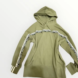 Olivgrön Adidas hoodie med logoband - Olivgrön hoodie från Adidas med huva och snörning. Snygga logoband med Adidas-tryck längs ärmarna och över bröstet. Mjukt material och klassiska ränder på mudden. Perfekt för en sportig och avslappnad stil.