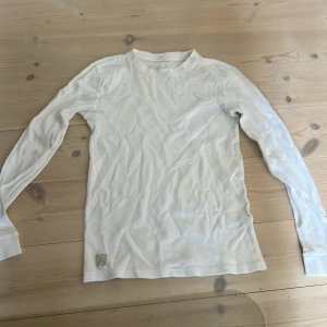 Vit långärmad tröja i bomull - En basic vit långärmad tröja i mjuk bomull. Tröjan har rund halsringning och en liten patch nertill på framsidan. Perfekt att styla lager på lager eller bära som den är. Enkel och clean look som funkar till det mesta.