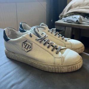Philipp Plein vita sneakers strl 44 - Snygga vita sneakers från Philipp Plein med svart detalj på hälen och tydlig logga på sidan. Skorna har snörning med märkta skosnören och präglad text runt sulan. Tillverkade i skinn för en lyxig känsla och passar perfekt till streetwear eller casual outfits.