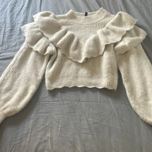 Beige stickad tröja med volang - Superfin beige stickad tröja med breda volanger över axlarna och bröstet. Tröjan har långärmade puffärmar och en croppad passform. Perfekt för dig som vill ha något gulligt och trendigt till hösten eller vintern💕