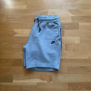 Grå Nike tech fleece shorts  - Snygga grå shorts från Nike med svart logga på ena benet. De har elastisk midja med svart snörning och praktiska fickor, varav en med dragkedja. Perfekta för träning eller chill. Mjukt bomullsmaterial för maximal komfort.