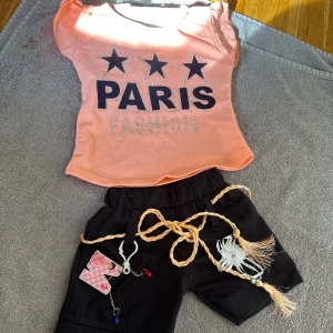 Persikofärgad t-shirt med Paris-tryck - Snygg persikofärgad t-shirt med korta ärmar och stort tryck med texten 'PARIS FASHION' och tre svarta stjärnor framtill. T-shirten har glittriga detaljer och är i mjuk bomull, perfekt för en trendig look.