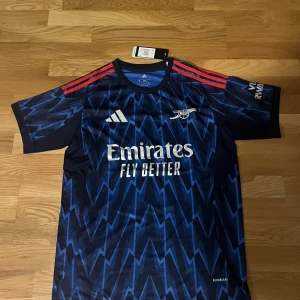 Arsenal bortatröja 25/26 - Arsenal bortatröja från Adidas i mörkblått med blixtmönster i blå toner. Tröjan har röda ränder på axlarna, klubbmärke och sponsortryck framtill samt 'Visit Rwanda' på ärmen. Tillverkad i lätt och ventilerande polyester.