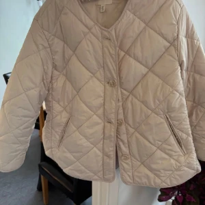 Beige quiltad jacka från H&M - Quiltad beige jacka från H&M i storlek S. Jackan har knappar och två fickor. Perfekt för lager-på-lager och har en stilren look med diskret glansigt tyg. Enkel att matcha till olika outfits.