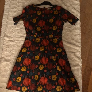 Blommig klänning från Cissi och Selma - Färgglad klänning från Cissi och Selma, modell Irma, med blommigt mönster i rött, gult och blått på mörk botten. Kort ärm, rund hals och dragkedja bak. Tillverkad i bomull med lite stretch och nederdelen är fodrad med polyester. Perfekt för dig som gillar retrostil och unika prints. Jättebra skick. Två fickor 
