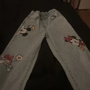 Ljusblå jeans med Disney-tryck - Unika ljusblå jeans med coola tryck av Minnie Mouse och Daisy Duck på benen. Byxorna har en loose passform och resår i midjan för extra komfort. Perfekta för dig som vill sticka ut med en lekfull och trendig stil.