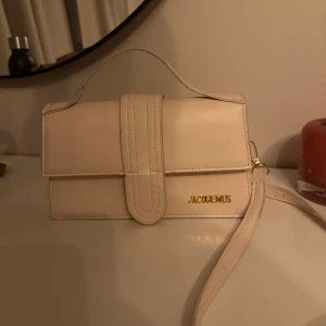 Beige Jacquemus axelväska i skinn - Säljer en stilren beige axelväska från Jacquemus i slätt skinn. Väskan har ett elegant handtag, avtagbar axelrem och guldiga detaljer med logga framtill. Perfekt storlek för det viktigaste och en clean look som lyfter varje outfit. Köpt på plick och fick inget äkthetsbevis därav billigare.