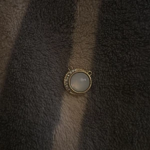 Guldigt hänge med vit sten och strass - Runt hänge i guldton med en mjölkvit sten i mitten och glittriga strassdetaljer på ena sidan. Perfekt att använda som berlock till halsband eller armband. Enkel men ändå lyxig design som ger en elegant touch.