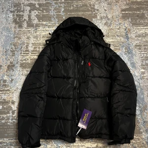 Svart pufferjacka från Polo Ralph Lauren - Svart pufferjacka från Polo Ralph Lauren med huva och dragkedja. Jackan har quiltad design, två sidofickor med dragkedja och en röd broderad logga på bröstet. Perfekt för kalla dagar och riktigt snygg till streetwear-stilen.