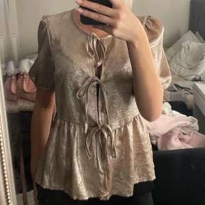 Champagnefärgad blus med knyt - Säljer en glansig champagnefärgad blus med puffärmar och tre dekorativa knyt framtill. Blusen har volang nedtill och är tillverkad i ett skimrande material som ger en lyxig känsla. Perfekt för dig som vill sticka ut med en trendig och feminin look. Inga defekter och nypris 179kr!