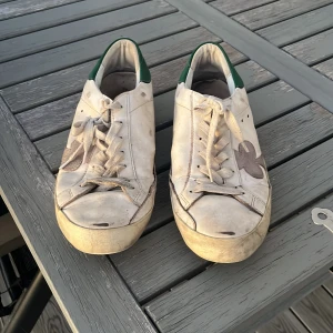 Vita sneakers The Gallerist med grön häl - Snygga vita sneakers från The Gallerist med gröna detaljer på hälen och ett stort klubbmärke på sidan. Skorna har snörning och platt sula, samt en unik svartvit detalj baktill. Perfekta för dig som gillar streetstyle och vill sticka ut.