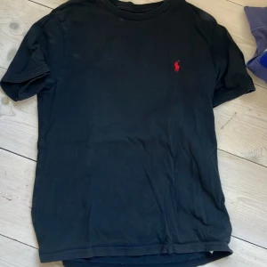 Svart t-shirt från Ralph Lauren, strl M 10-12 - Svart t-shirt från Ralph Lauren med klassisk röd broderad logga på bröstet. Enkel och stilren design med rund hals och korta ärmar. Perfekt till jeans eller shorts för en clean look.