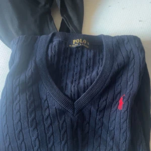 Mörkblå kabelstickad tröja Polo Ralph Lauren - Kabelstickad mörkblå tröja från Polo Ralph Lauren med röd broderad logga på bröstet. V-ringad modell med långa ärmar och ribbade muddar. Klassisk och stilren design i mjuk bomull, perfekt för lager-på-lager under höst och vinter.