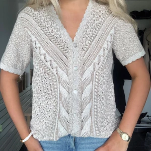 Beige stickad kofta med kort ärm - Superfin beige stickad kofta med korta ärmar och snyggt mönster. Koftan har v-ringning och knäpps med pärlemorknappar framtill. Perfekt till jeans för en chill och trendig look.