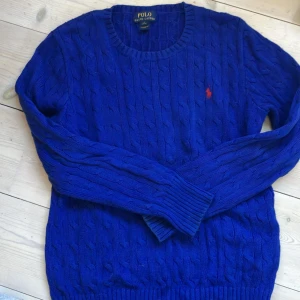 Blå kabelstickad tröja Polo Ralph Lauren, strl L 14-16 - Kabelstickad blå tröja från Polo Ralph Lauren med rund halsringning och klassisk röd logga broderad på bröstet. 