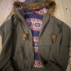 Grå hoodiejacka med fuskpäls och mönster - Mysig grå hoodiejacka med dragkedja, fuskpäls runt huvan och snörning med pälsbollar. Insidan har ett blått, rött och vitt vintermönster. Perfekt för kalla dagar när du vill vara både varm och snygg.