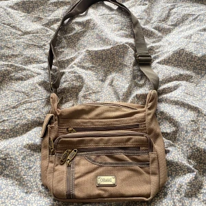 Beige axelväska från OR&MI i bomull - Säljer en beige axelväska från OR&MI med flera praktiska fack och dragkedjor. Väskan är tillverkad i slitstark bomull och har justerbar axelrem. Perfekt för dig som vill ha en stilren och funktionell väska till vardagen.