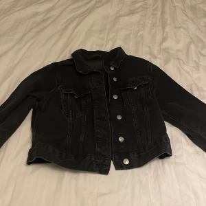 Svart jeansjacka med silvriga knappar - Snygg svart jeansjacka med klassisk krage och silvriga knappar framtill. Jackan har två bröstfickor med lock och knapp, samt detaljerade sömmar för en cool look. Perfekt att slänga över en hoodie eller t-shirt.