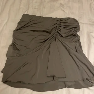 Grå draperad kortkjol med rynk - Snygg grå kortkjol med draperad design och rynkade detaljer på sidorna. Kjolen har en asymmetrisk nederkant och är tillverkad i ett mjukt, lätt material som ger ett fint fall. Perfekt för dig som gillar en trendig och stilren look. 