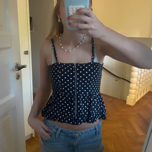 Svart prickig croppad linnetopp - Supersöt svart croppad linnetopp med vita prickar och volang nertill. Toppen har smala axelband och dragkedja framtill som detalj. Perfekt för varma dagar och ger en lekfull vibe till outfiten.