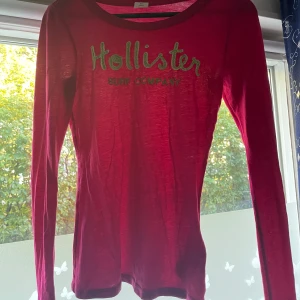 Röd långärmad tröja från Hollister - Snygg röd långärmad tröja från Hollister med trycket 'Hollister Surf Company' i grönt framtill. Tröjan har rund hals och är gjord i mjuk bomull. Passformen är normal/lite stor och materialet är lätt och skönt.