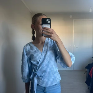 Blåvit omlottblus - Supersöt blåvit rutig omlottblus från Samsa med knyt i midjan. Blusen har v-ringning och har puffiga ärmar med resår vid ärmslut. 