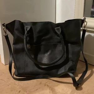 Svart axelväska i skinnimitation - Snygg svart axelväska med både handtag och justerbar axelrem. Väskan har enkel design med en ficka framtill och detaljer i guld. Perfekt storlek för att få plats med allt du behöver. Stilren och passar till många outfits.