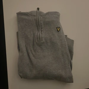 Grå half zip tröja från Lyle & Scott - Säljer en stilren grå långärmad tröja från Lyle & Scott Junior med klassisk gul logga på bröstet. Tröjan har half zip-dragkedja och är i mjukt material. 