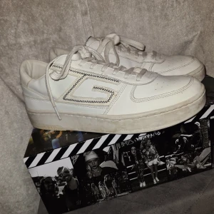 Vita sneakers från Guess, strl 43 - Snygga vita sneakers från Guess med klassisk låg profil och coolt sicksackmönster runt det stora G:et på sidan. Skorna har snörning, perforerad tå och är gjorda i skinn. Perfekta för dig som gillar stilrena och trendiga sneakers.