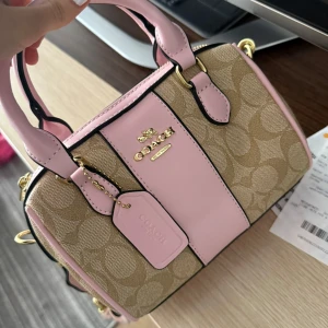 Coach axelväska i rosa och beige canvas - Superfin axelväska från Coach i beige och rosa canvas med klassiskt logomönster. Väskan har detaljer i ljusrosa skinn, guldfärgade metalldetaljer och bred axelrem med Coach-tryck. Perfekt storlek för det viktigaste och riktigt snygg att bära crossbody.