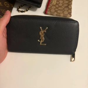 Svart YSL plånbok i skinn med dragkedja - Snygg svart plånbok från YSL i äkta skinn med guldigt YSL-logo framtill. Plånboken har dragkedja runt om och stilren design som passar perfekt i väskan. Lyxig känsla och klassisk look.