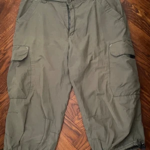 Olivgröna cargoshorts med snörning - Säljer ett par olivgröna cargoshorts med flera stora fickor på sidorna och baktill. Shortsen har snörning i bensluten och dragkedja framtill. Perfekta för dig som gillar praktiska detaljer och en loose fit. Materialet känns som bomull eller polyester.