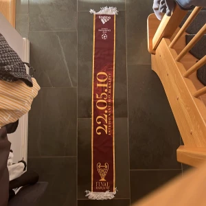 Authentic Champions League Final halsduk - Säljer en exklusiv halsduk från Champions League-finalen 2010 i Madrid. Halsduken är vinröd med gula detaljer och text, har vita fransar i ändarna och Adidas-logga upptill. Perfekt för dig som älskar fotboll och samlar på unika supporterprylar.