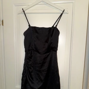 Svart satin klänning med smala axelband - Supersnygg svart kort klänning från Bik Bok i glansigt satinmaterial. Den har smala justerbara axelband och en enkel, stilren design med lätt rynkade detaljer i sidan. Perfekt för dig som vill ha en klassisk look med modern touch.