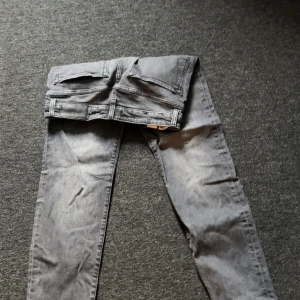 Grå jeans från BOSS, storlek 30/32 - Snygga grå jeans från BOSS med klassisk femficksdesign och svarta detaljer vid linningen. Jeansen har raka ben och subtila slitningar för en cool look. Tillverkade i bomull med en skön passform och diskret BOSS-logga på bakfickan.