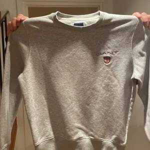 Grå sweatshirt från GANT i bomull - Klassisk grå sweatshirt från GANT med broderad logga och sköld på bröstet. Tröjan har rund halsringning, ribbade muddar och är tillverkad i mjuk bomull. Stl M.