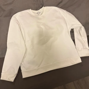 Vit sweatshirt med konstnärligt tryck - Vit sweatshirt med rund hals och långa ärmar. På ryggen finns ett stort konstnärligt tryck i svart och brunt som föreställer abstrakta figurer. Tröjan har en clean look framtill och är tillverkad i mjuk bomull för en skön känsla.