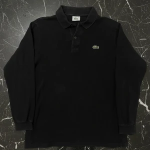 Svart/grå långärmad pikétröja Lacoste - Klassisk svart/grå långärmad pikétröja från Lacoste med ikoniska krokodilloggan på bröstet. Tröjan har krage, knappar vid halsen och är tillverkad i bomull. Perfekt för en clean och stilren look.