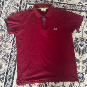 Vinröd pikétröja från Lacoste - Vinröd pikétröja från Lacoste med klassisk krage och dragkedja framtill. Tröjan har korta ärmar med mörkblå detaljer vid ärmslut och dragkedja. Ikoniska gröna krokodilloggan på bröstet. Tillverkad i mjuk bomull, perfekt för en clean och sportig look.