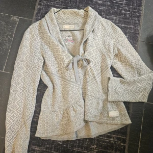 Gråmönstrad kofta från Odd Molly - Supersnygg grå och vit kofta från Odd Molly med stickat mönster och broderad krona på ryggen. Koftan har knytband framtill, lång ärm och en feminin passform. Perfekt för lager-på-lager och att lyfta din outfit.