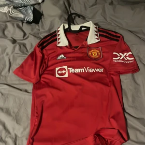 Manchester United Adidas matchtröja röd - Manchester Uniteds officiella matchtröja från Adidas i rött med vita och svarta detaljer. Klassisk krage med mönster, klubbmärke broderat på bröstet och TeamViewer-logga. Tillverkad i lätt och ventilerande polyester, perfekt för fotboll eller supporterliv.