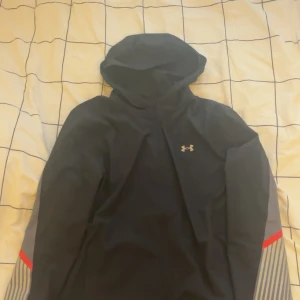 Svart vindjacka från Under Armour - Svart vindjacka från Under Armour med huva och dragkedja. Jackan har grå och röda detaljer på ärmarna samt diskret logga på bröstet. Tillverkad i polyester och har en sportig, fitted passform. Perfekt för träning eller utomhusaktiviteter.