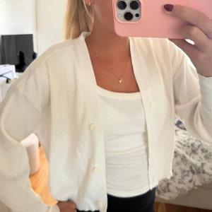 Vit kofta  - Säljer en fräsch vit kofta med croppad passform och stora knappar framtill. Koftan har långärmade ärmar och är stickad i mjukt material. Enkel och clean look som funkar till många stilar.