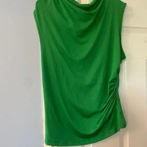 Grön ärmlös topp med rynkdetalj - Snygg grön ärmlös topp med draperad halsringning och rynk på ena sidan som ger en cool twist. Mjuk och stretchig känsla, perfekt för dig som gillar färg och vill sticka ut lite extra. Passar till både jeans och kjol.