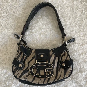 Zebramönstrad axelväska från Guess - Vintage 2000-tals axelväska från Guess med zebramönster i svart och beige. Väskan är snygg och unik med sitt stora G-spänne framtill. Inuti är fodret mönstrat med Guess-loggor och det finns en innerficka med dragkedja. Defekter: små fläckar inuti, kom privat vid intresse av fler bilder på detta!❤️Ungefärliga mått: Bredd 24 cm, Höjd 13,5 cm