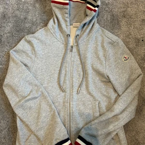 Grå Moncler hoodie med dragkedja - Snygg grå hoodie från Moncler med dragkedja, huva och snörning. Tröjan har Moncler-logga på ärmen och ribbade muddar med röd, vit och marinblå rand. Perfekt för chill dagar och streetstyle. Skick 9/10 skriv vid funderingar 