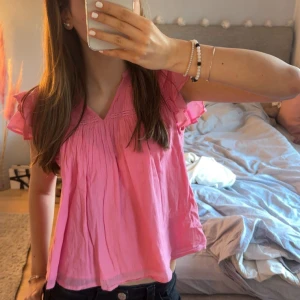 Rosa blus med volangärmar - Supersöt rosa blus med v-ringning och volangärmar. Blusen har en luftig och flowy passform, fina rynkade detaljer upptill och är tillverkad i ett tunt, lätt tyg. Perfekt för dig som gillar en feminin och avslappnad stil.