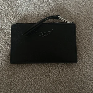 Zadig plånbok - Snygg svart plånbok i skinn med silverfärgad dragkedja och ett stilrent vingmotiv på framsidan. Praktisk storlek med dragkedja runt om och en smidig rem för att hålla i. Perfekt för kort, mynt och småsaker. Lite sliten bara vid märket annars brs